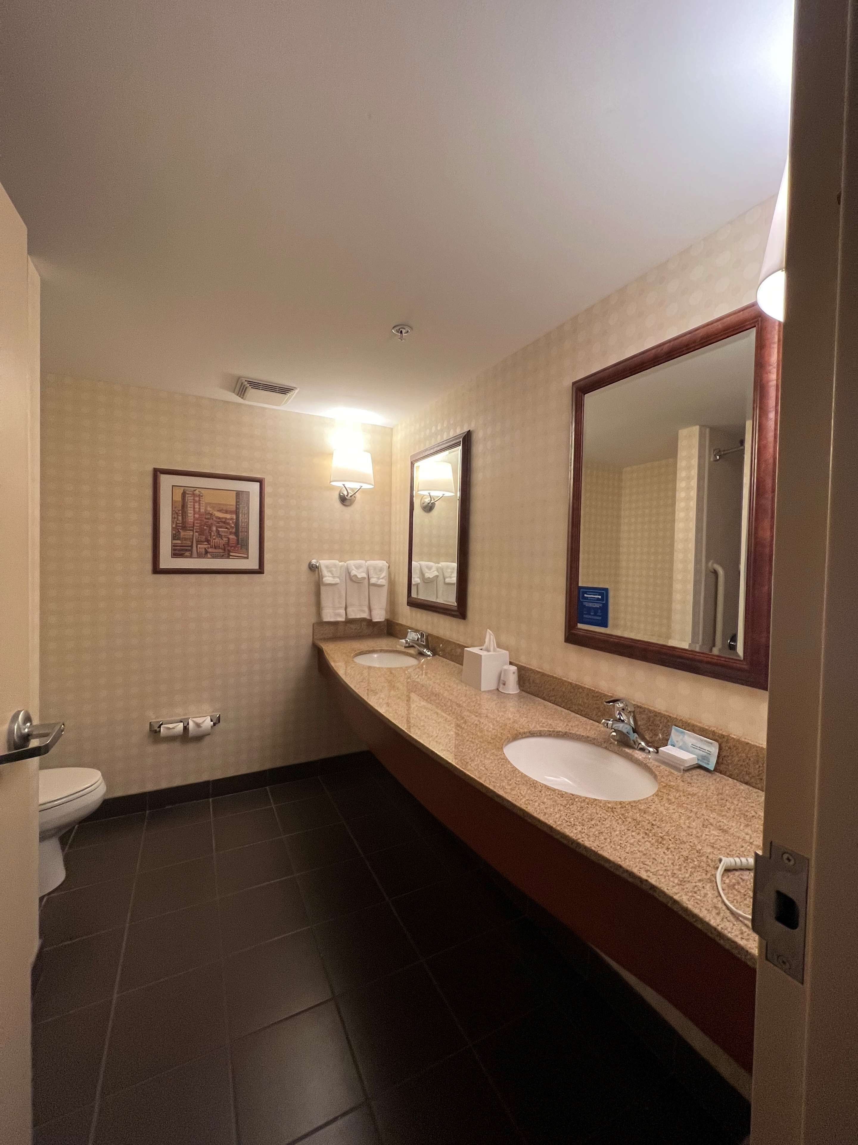 A hotel bathroom with two sinks, a large mirror, a framed picture on the wall, and a toilet.