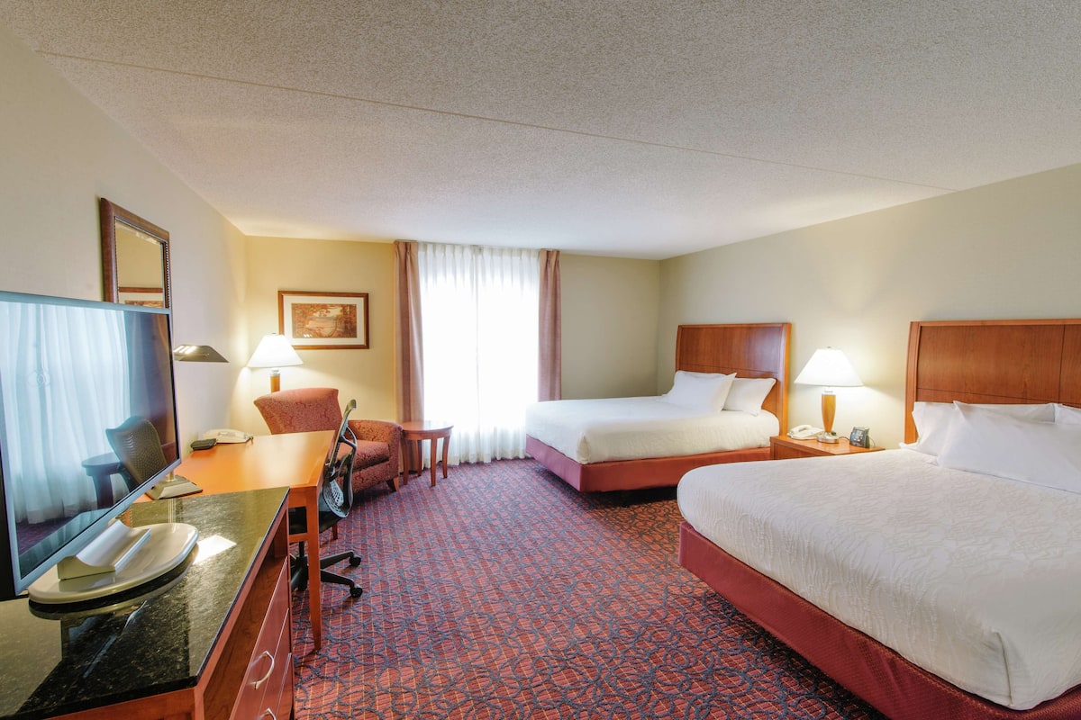 A hotel room with two beds, a desk with a computer, a chair, a small table, and a window with curtains.