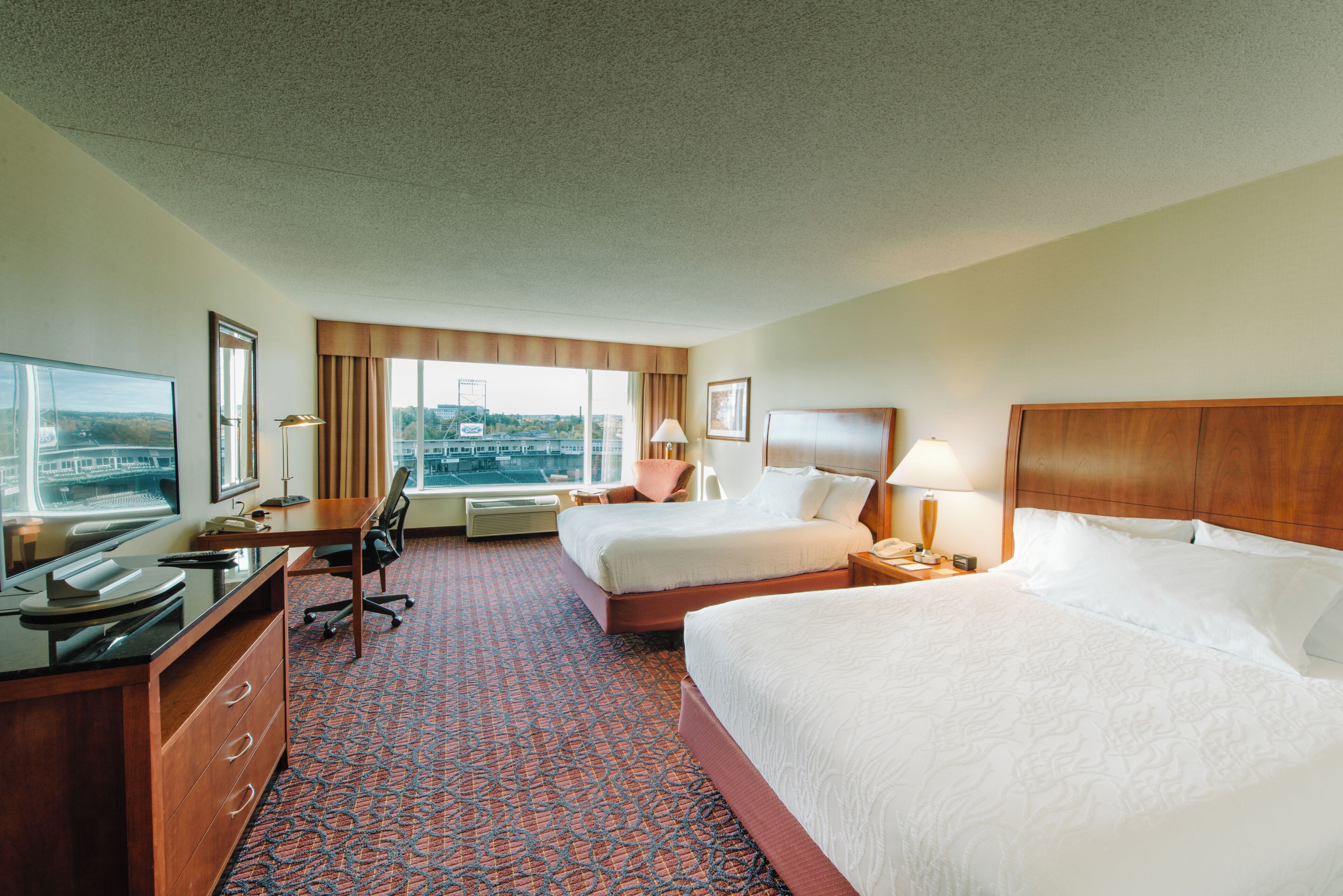 A hotel room with two beds, a desk, a chair, a TV, and a window with a city view.