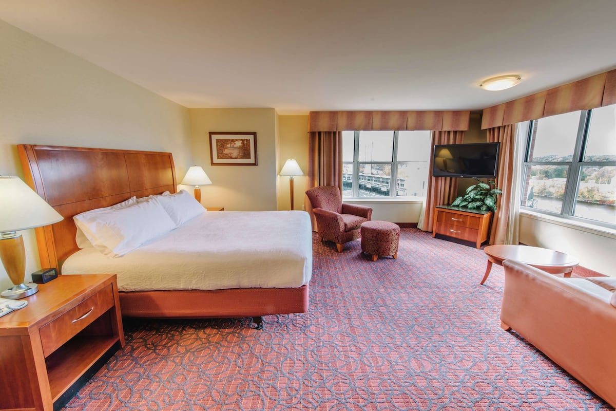 A hotel room with a large bed, two chairs, a small table, a TV, and a window with a city view.