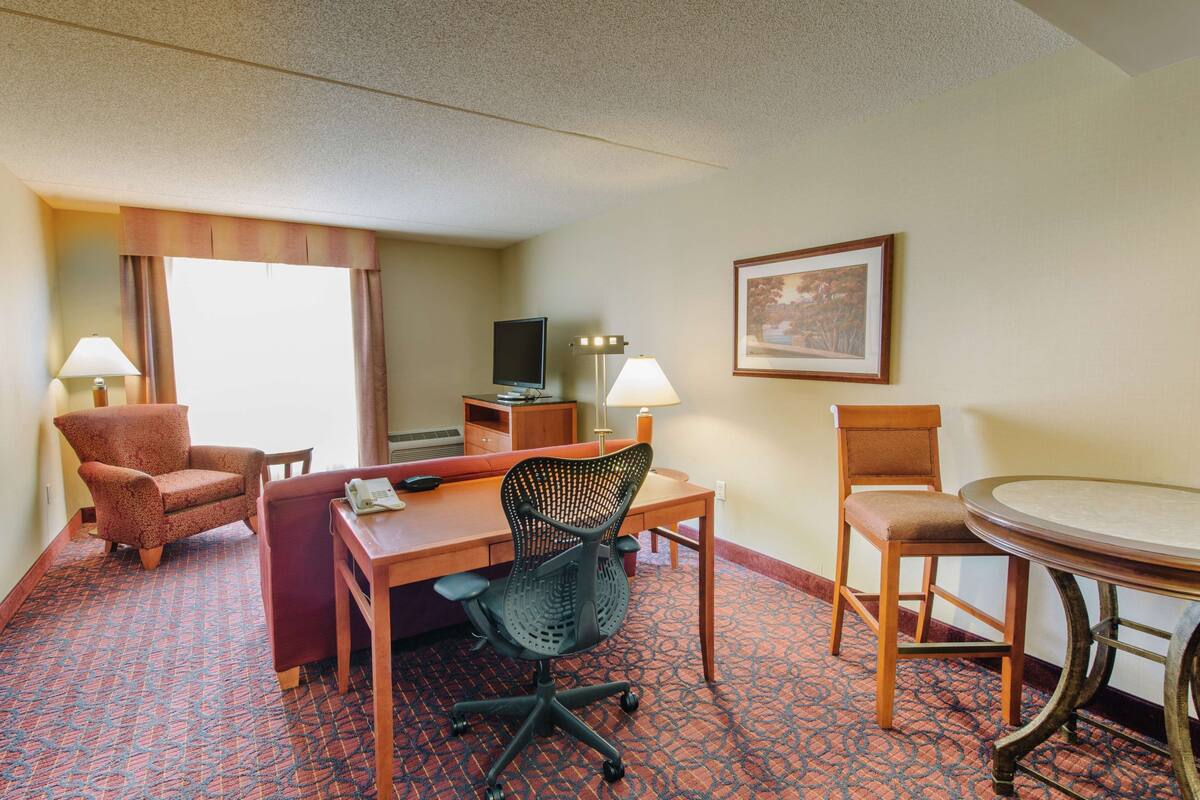 A hotel room with a desk, two chairs, a television, and a painting on the wall.