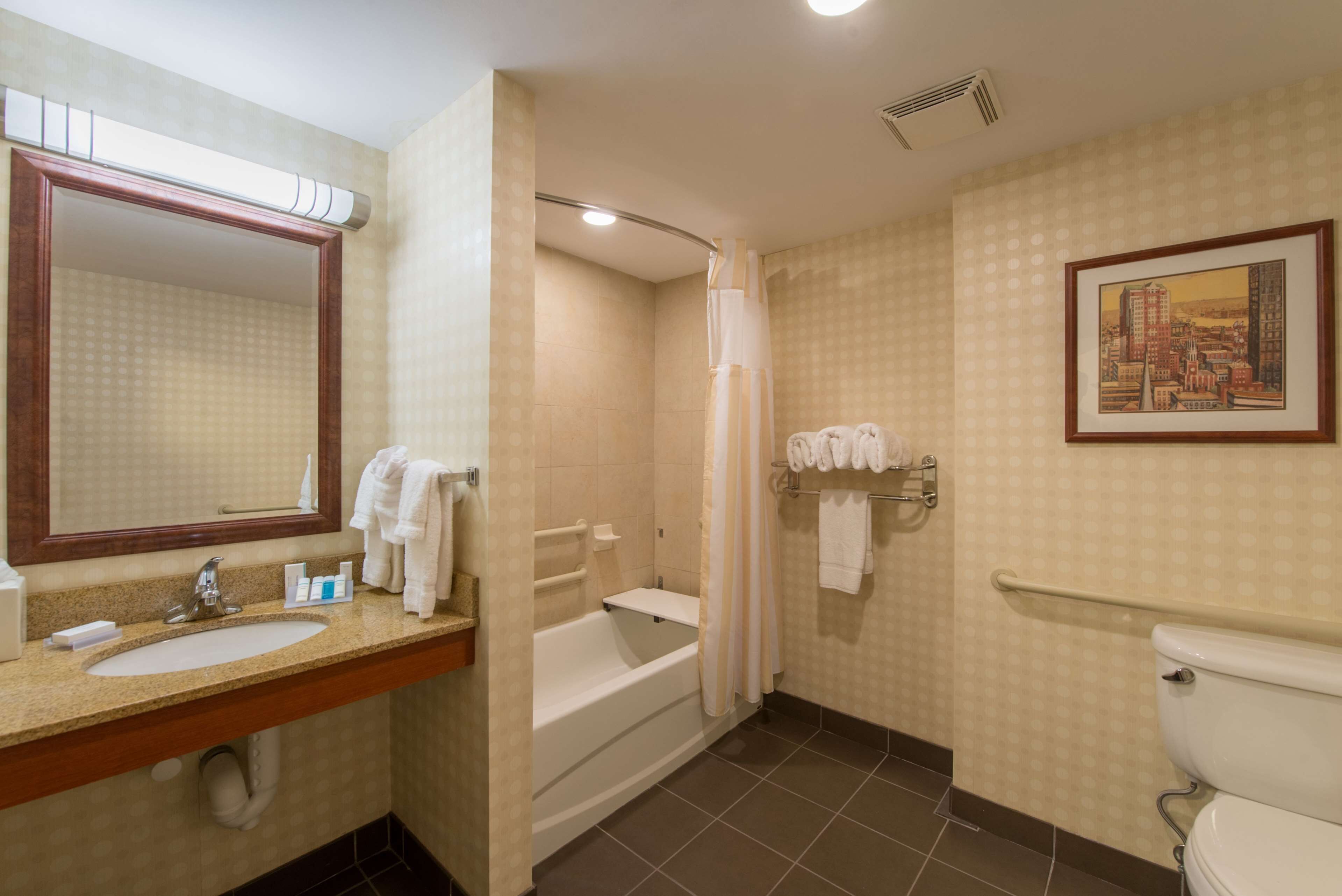 A hotel bathroom with a sink, mirror, bathtub, shower, toilet, and a framed picture on the wall.