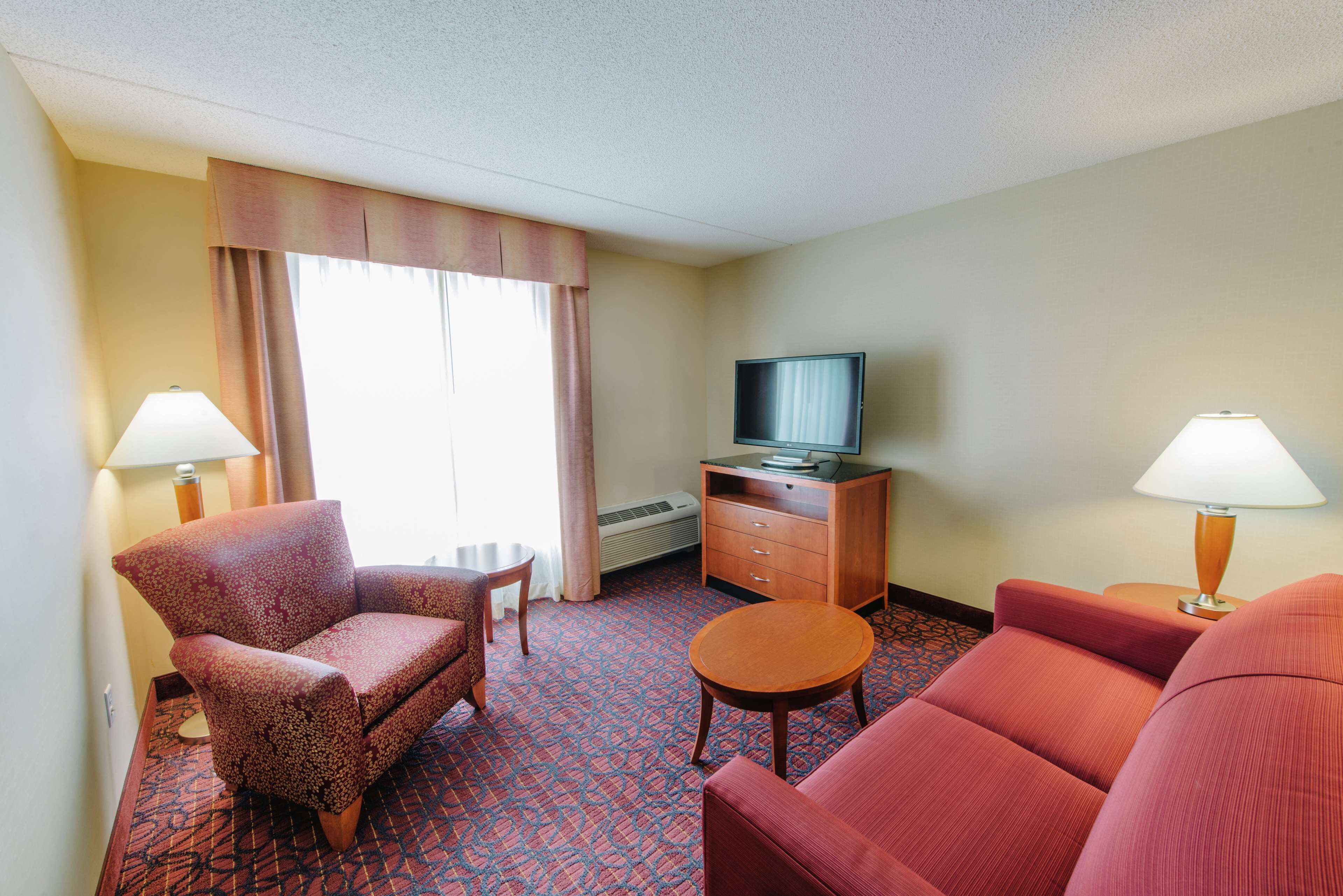 A hotel room with a red carpet, a television on a wooden cabinet, two lamps, and a large window with curtains.