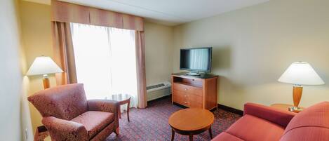 A hotel room with a red carpet, a television on a wooden cabinet, two lamps, and a large window with curtains.