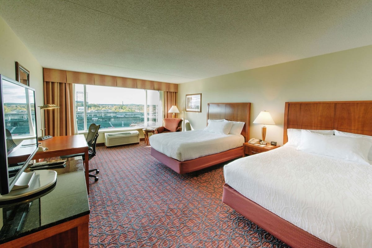 A hotel room with two beds, a desk, a chair, a TV, and a large window with a view.