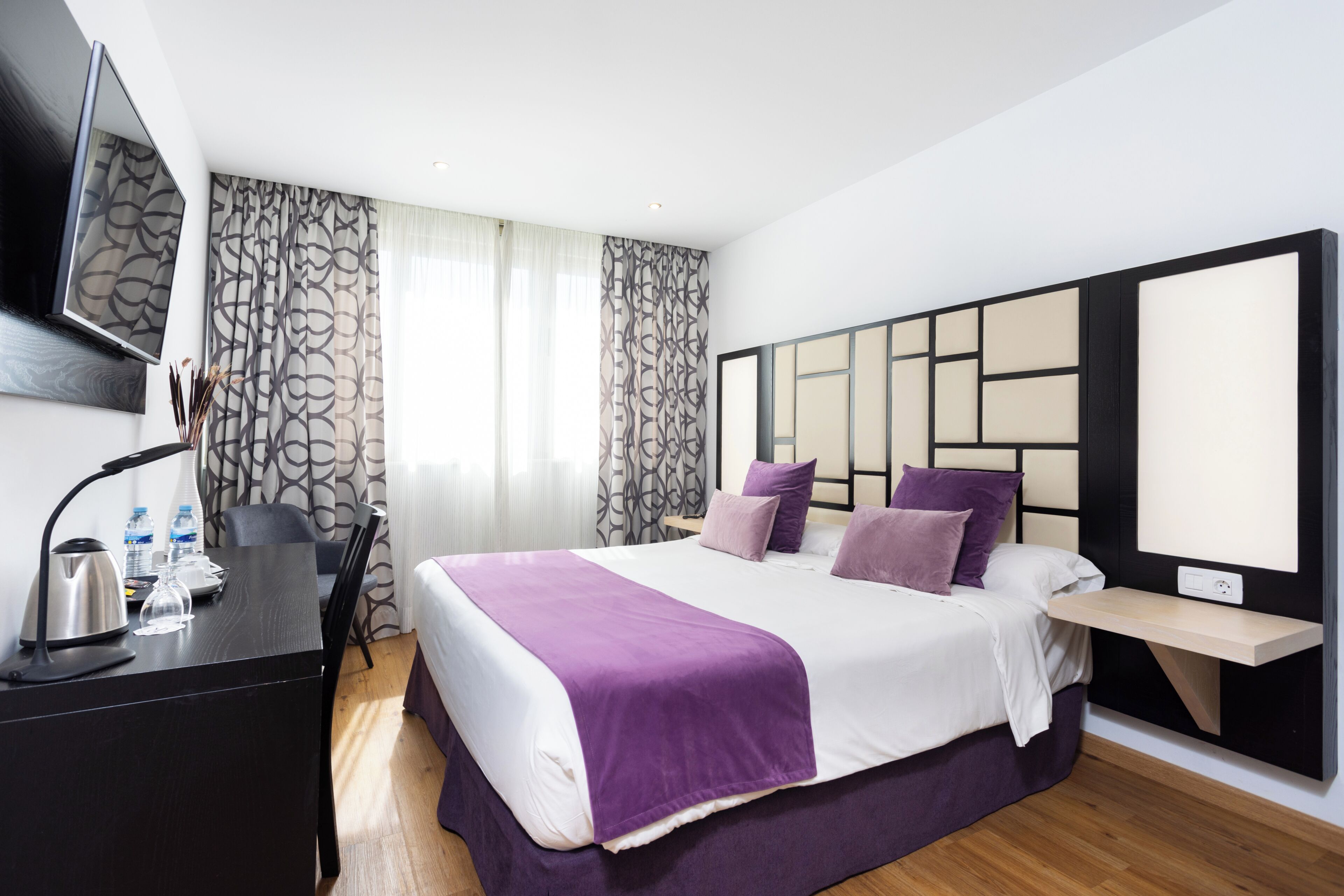 A modern hotel room with a large bed, a desk with a lamp, a flat-screen TV, and a view of the city.