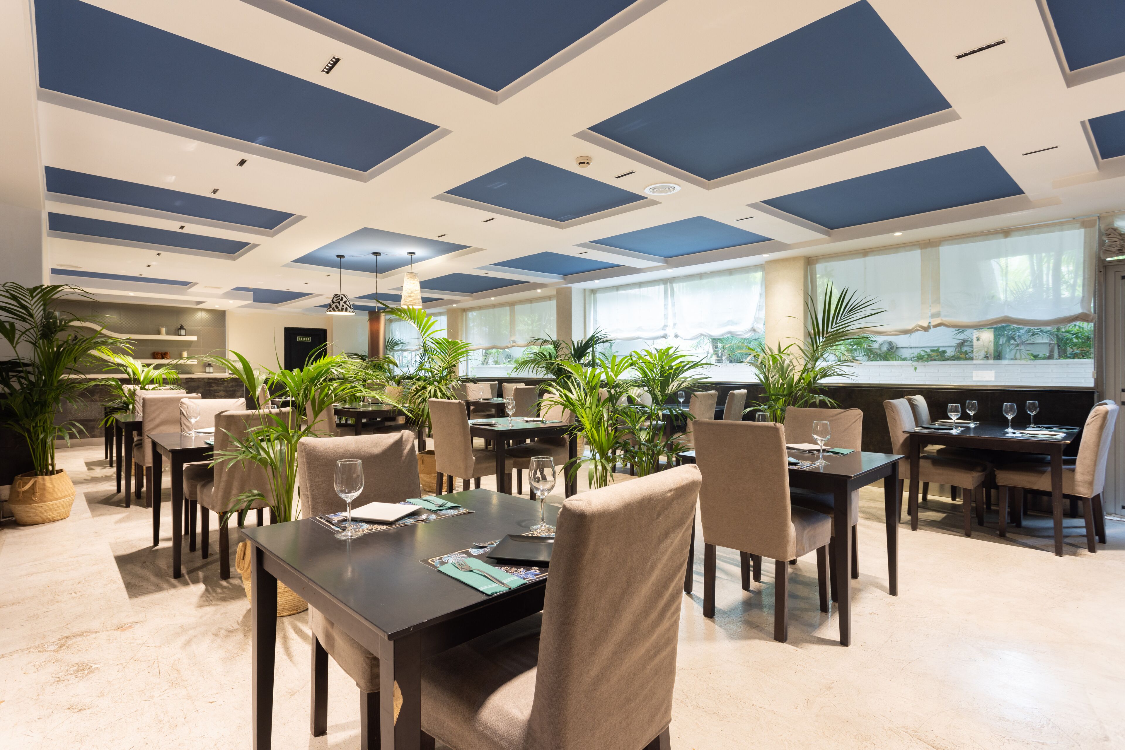 Un espace de restauration moderne, doté de plusieurs tables et chaises, d’un plafond orné de panneaux bleus et blancs, et de grandes fenêtres offrant une vue sur la verdure environnante.