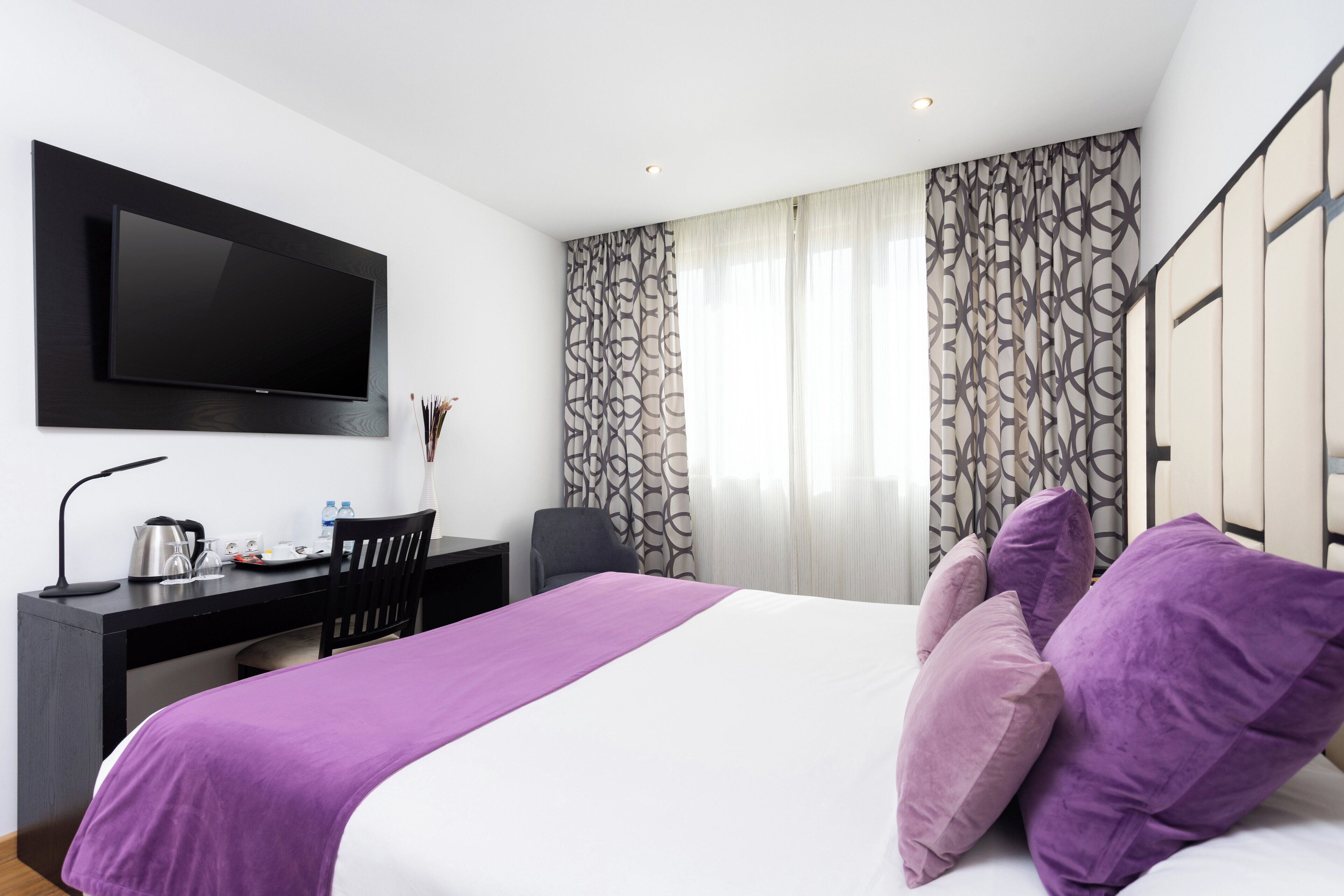 A modern hotel room with a large bed, a desk with a flat-screen TV, and a window with patterned curtains.