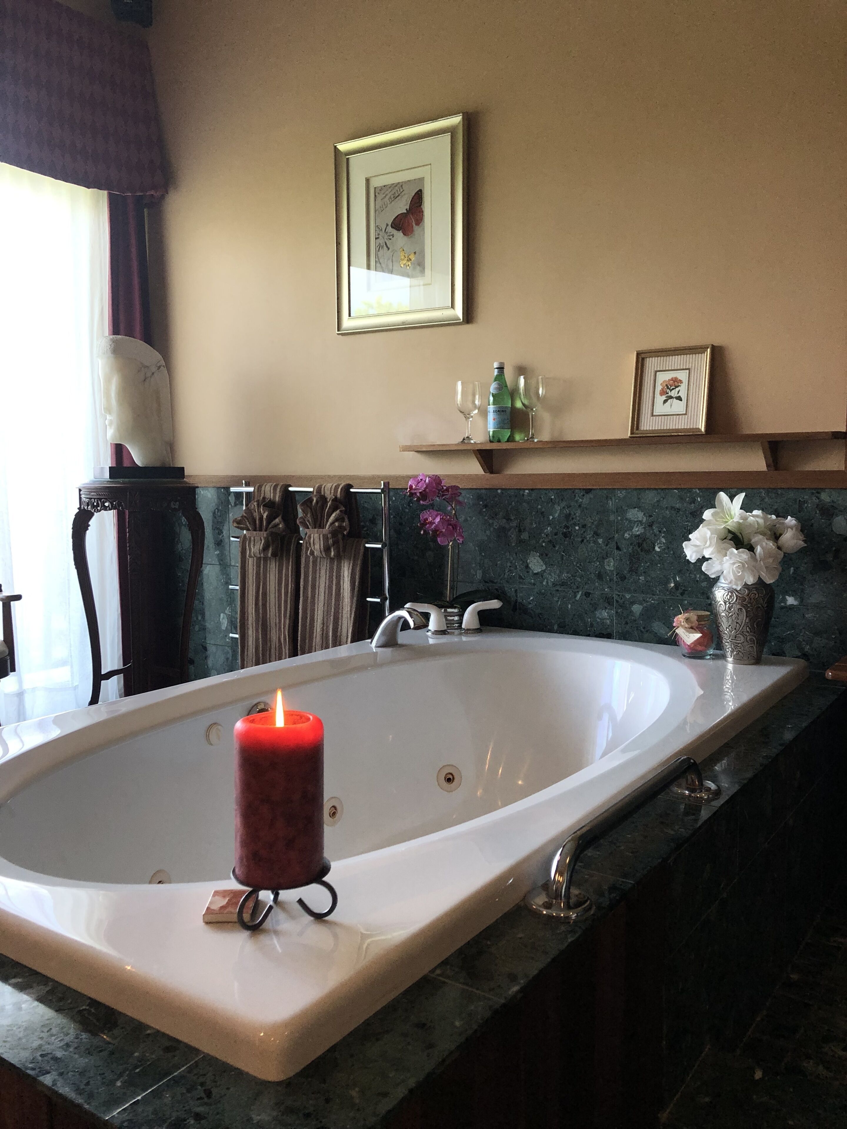 A bathroom with a large bathtub, a lit candle, a marble countertop, a towel rack, a framed picture on the wall, and a shelf with bottles and glasses.