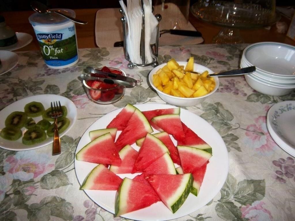 A dining table with plates of watermelon, kiwi, and pineapple slices, a container of plain yogurt, and a floral tablecloth.