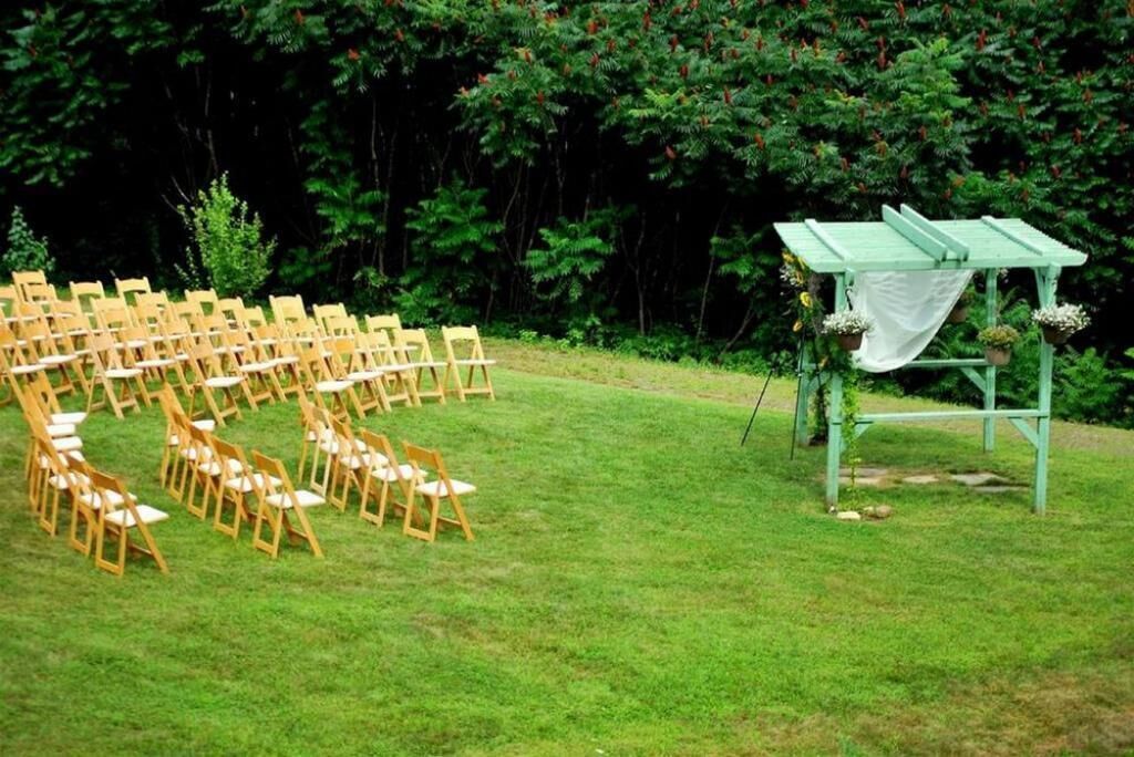 A grassy outdoor area with wooden chairs arranged in rows, a green wooden structure with potted plants, and dense green foliage in the background.