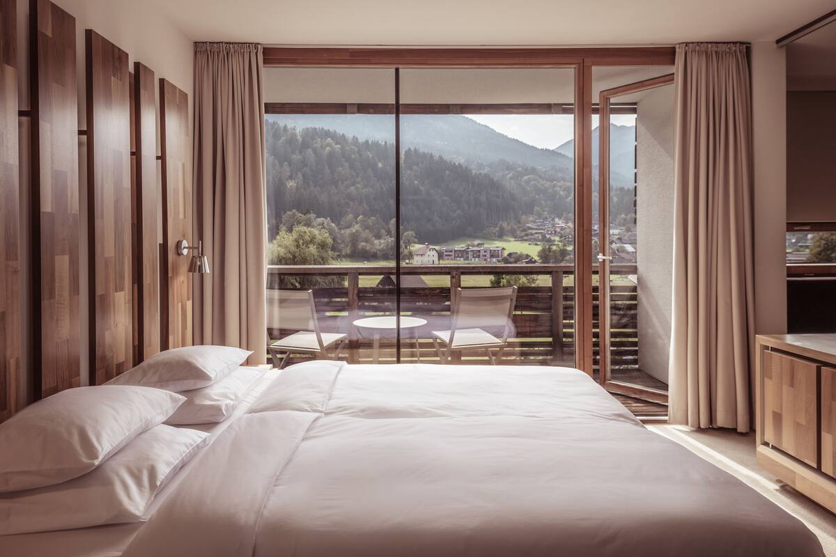 Un dormitorio con una cama grande, un balcón con mesa y sillas, y vista a un paisaje montañoso.