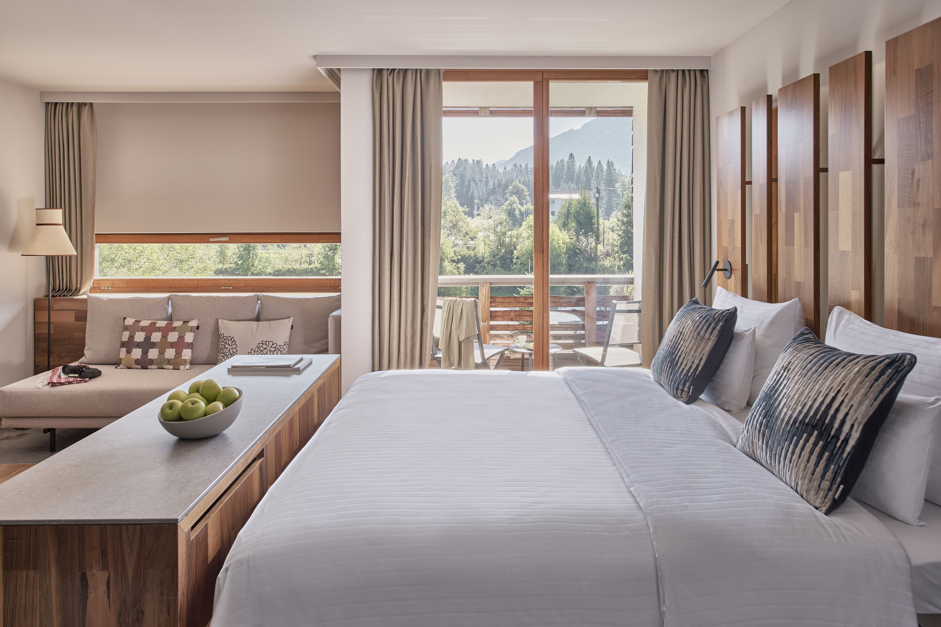 A hotel room with a large bed, a desk with a bowl of apples, a view of trees and mountains through the window, and a balcony with a wooden railing.
