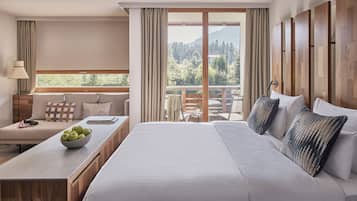 Une chambre d’hôtel avec un grand lit, un bureau sur lequel se trouve un panier de pommes, une vue sur les arbres et les montagnes par la fenêtre, et un balcon doté d’une balustrade en bois.