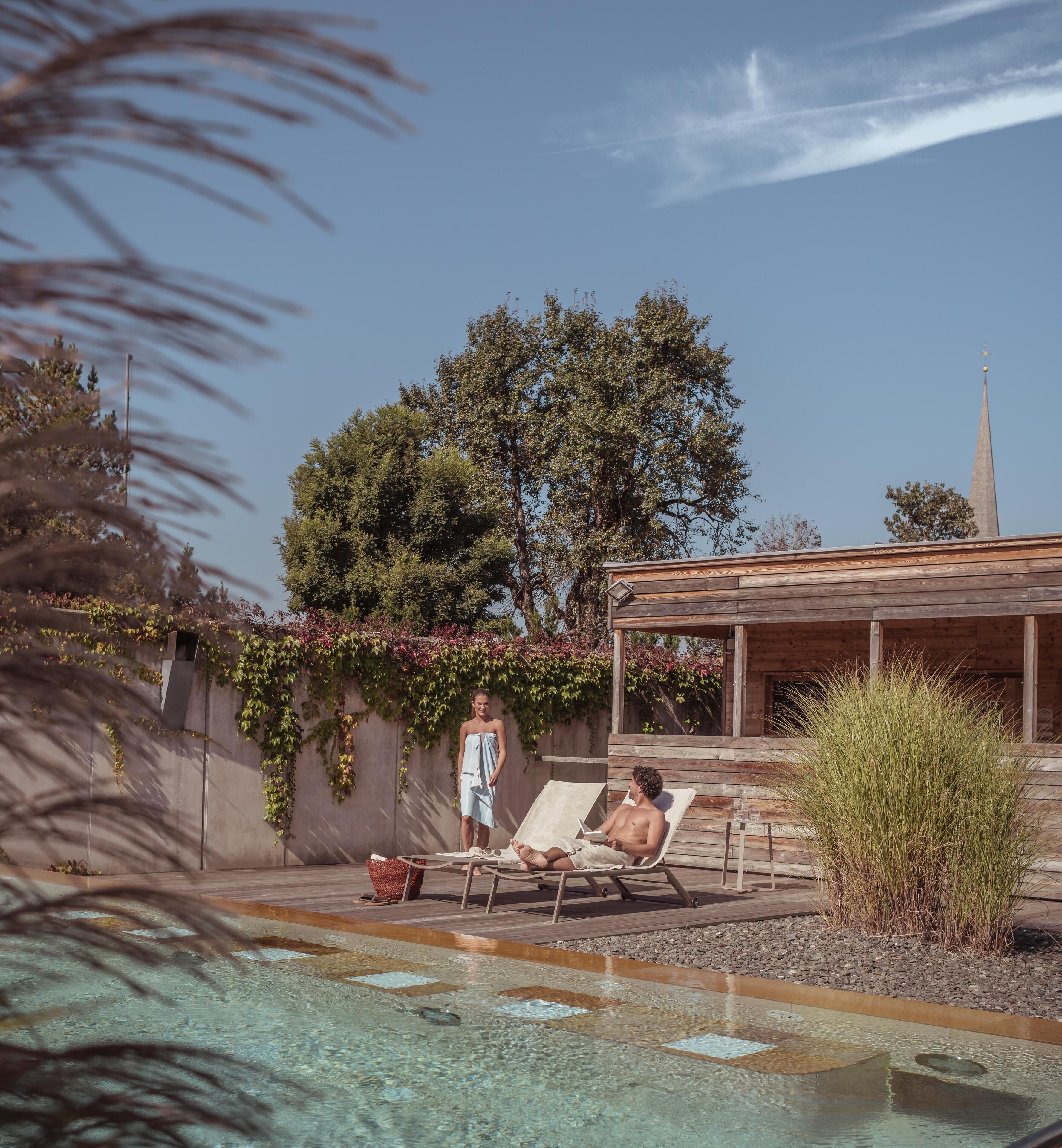 A pool area with lounge chairs, a wooden pergola, and a view of a church spire.