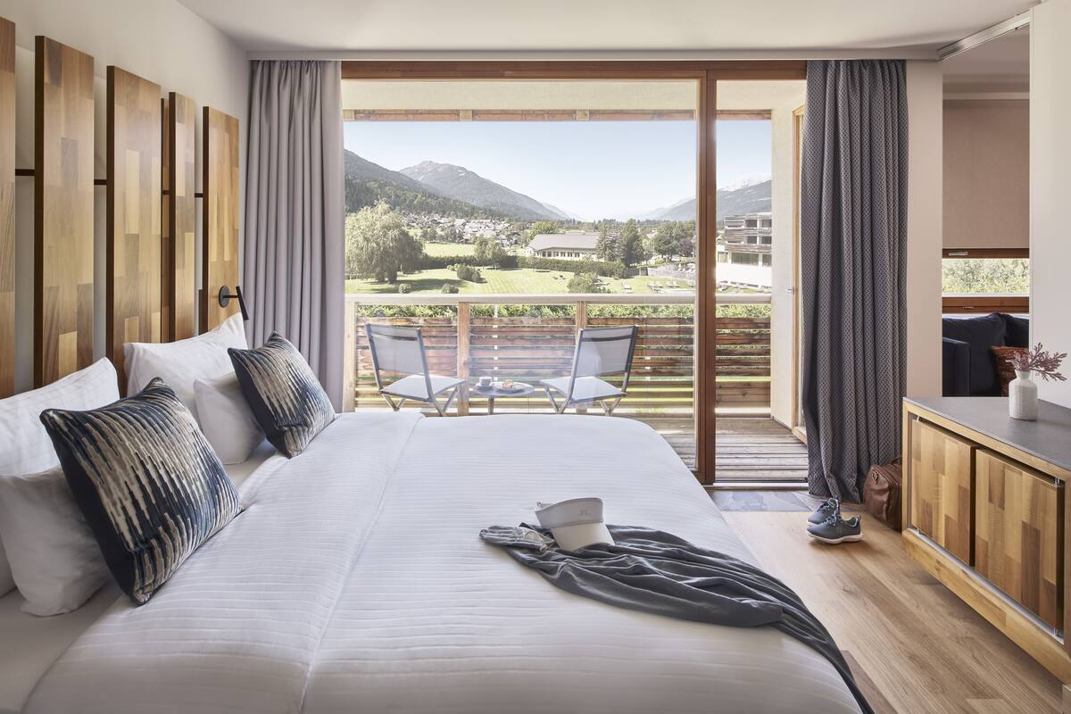 Habitación de hotel con una cama grande, un balcón con vistas a las montañas y un comedor con mesa y sillas.