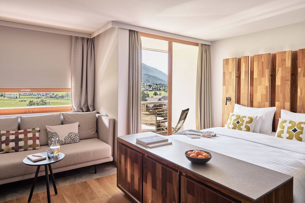 Habitación de hotel con una cama grande, cabecera de madera, un sofá, una mesita y vistas a un paisaje montañoso.