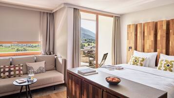 A hotel room with a large bed, a wooden headboard, a sofa, a small table, and a view of a mountainous landscape.
