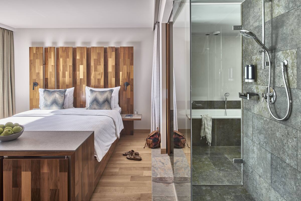 Una habitación de hotel moderna con cabecera de madera, una cama grande con ropa de cama blanca y una mesita de noche con un frutero, y una zona de ducha con paredes de cristal.