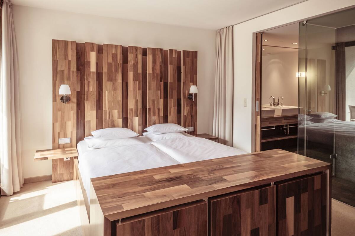 Una habitación de hotel moderna con cabecera de madera, una cama grande, mesitas de noche, un escritorio de madera y una zona de ducha de cristal.