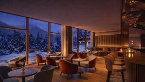 Ein modernes Interieur mit großen Fenstern und Blick auf verschneite Berge, gemütliche Sitzbereiche und eine Bar/Lounge mit warmer Beleuchtung.