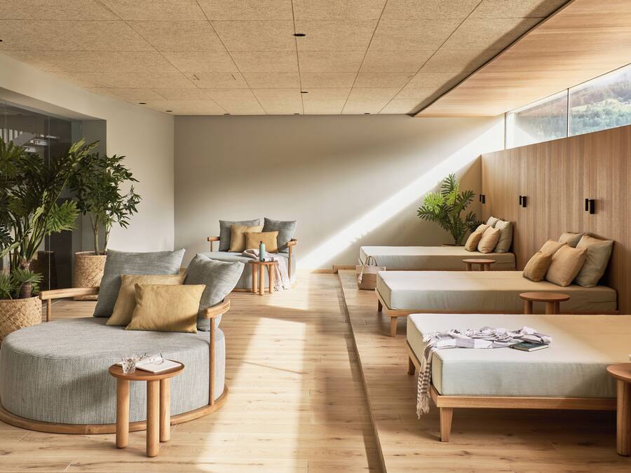 Una habitación moderna y luminosa, con piso de madera, un sofá, una silla y una mesita. Hay plantas en macetas y un gran ventanal que proporciona luz natural.