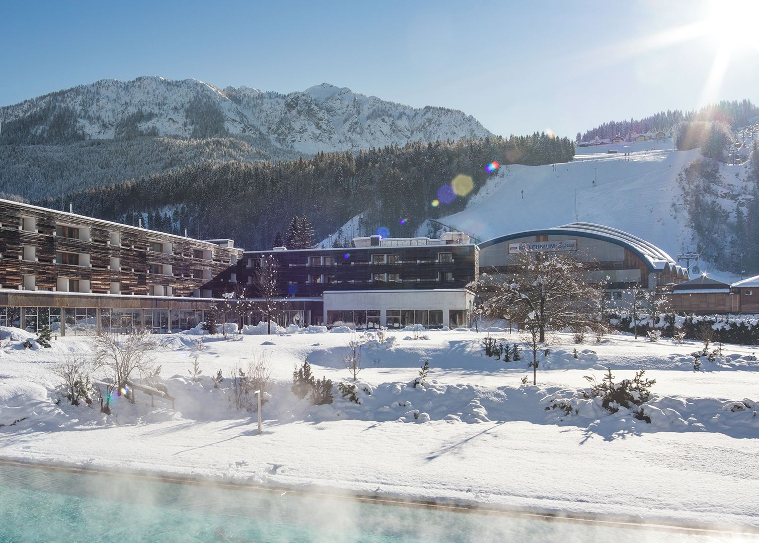 A modern building with a pool, surrounded by snow-covered mountains and a ski slope.
