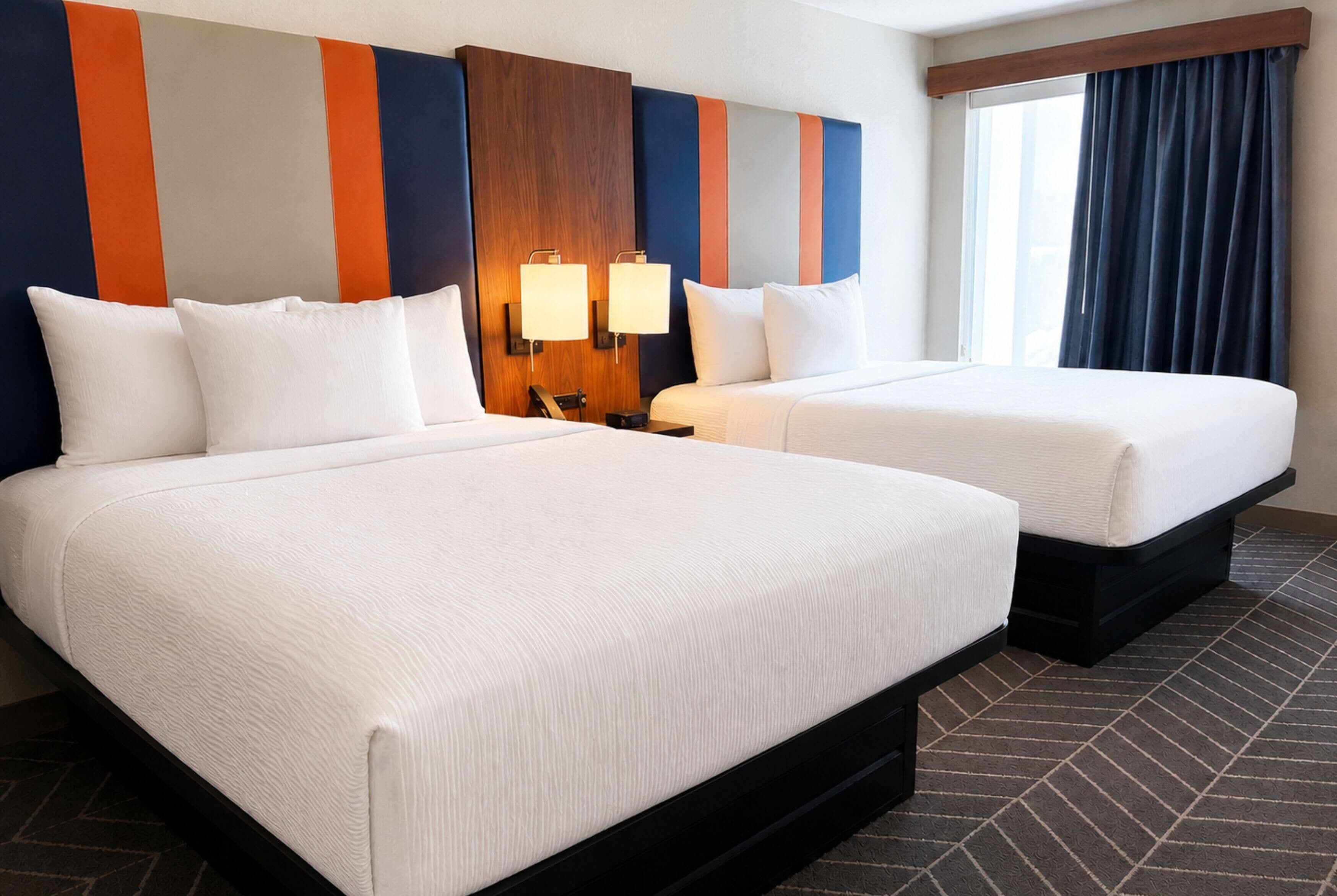 A hotel room with two beds, a wooden headboard, a nightstand, a lamp, and a window with curtains.