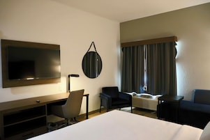 Habitación de hotel con cama, escritorio, silla, televisor, espejo y ventana con cortinas.