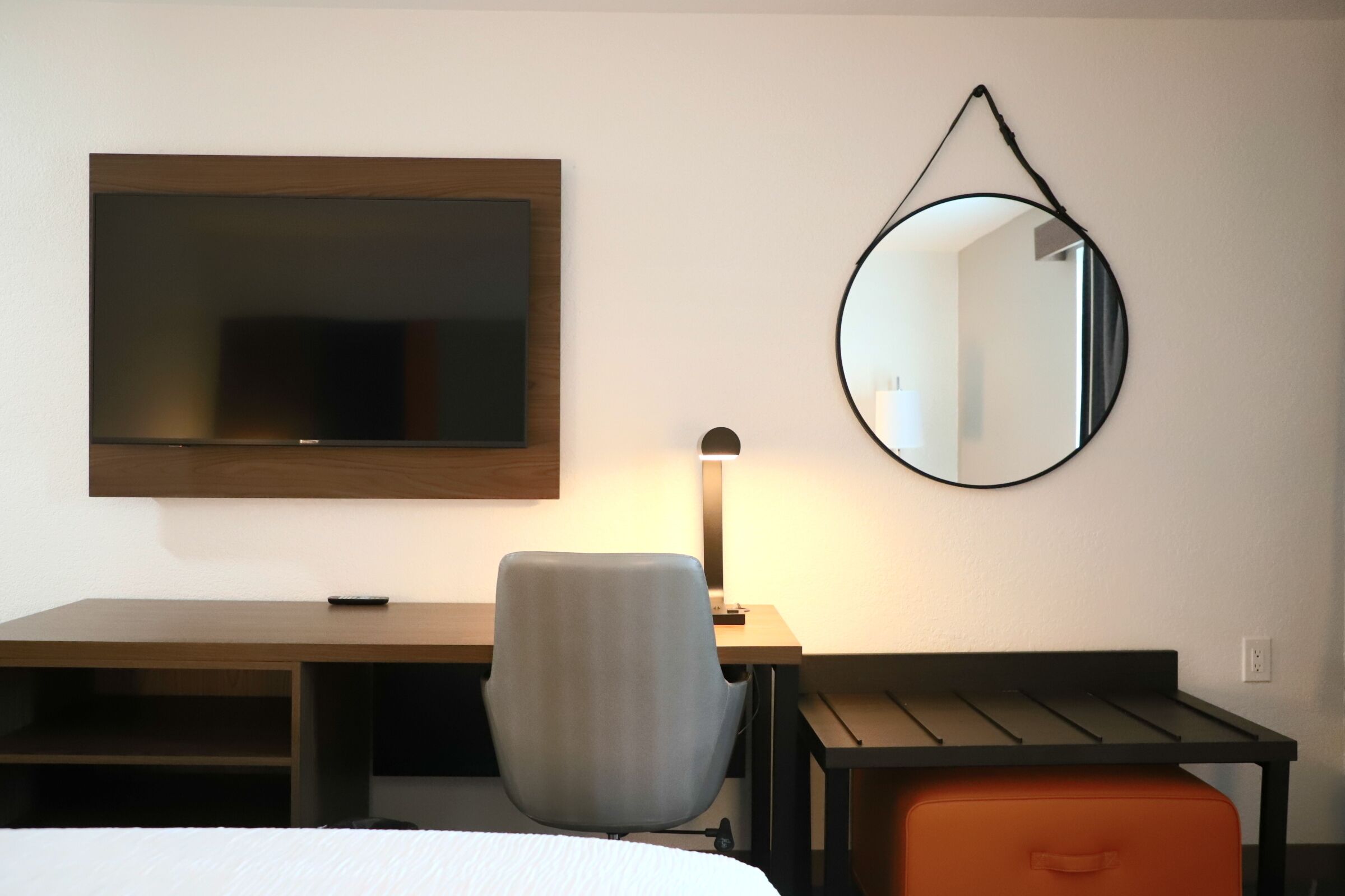 A hotel room with a bed, a desk, a chair, a desk lamp, a round mirror, and a small orange storage unit.