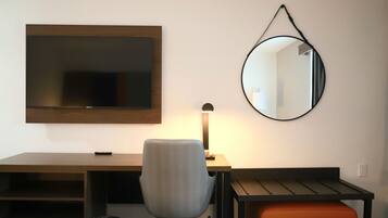 A hotel room with a bed, a desk, a chair, a desk lamp, a round mirror, and a small orange storage unit.