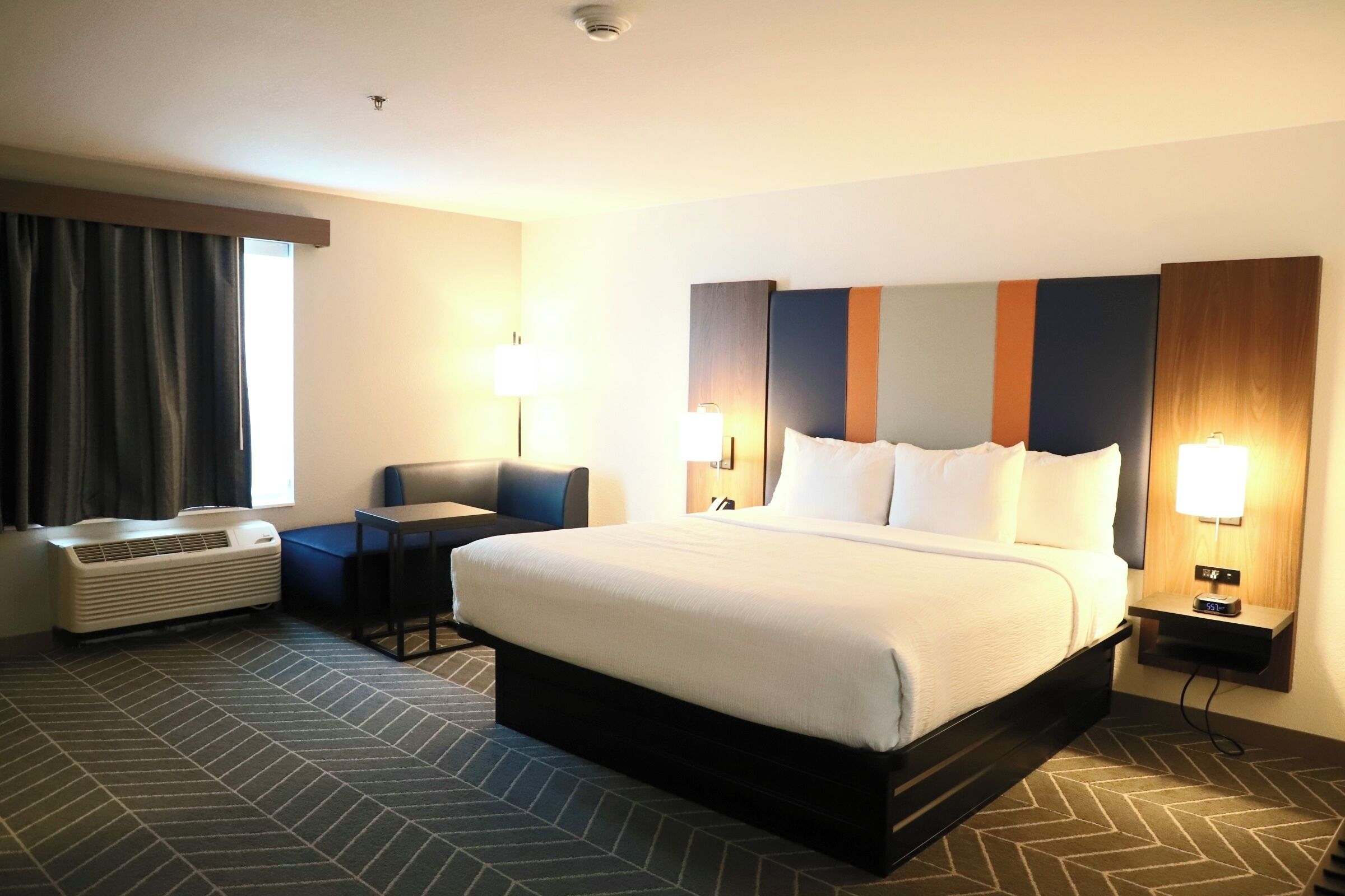 A hotel room with a bed, a desk, a chair, a heater, and a window with curtains.