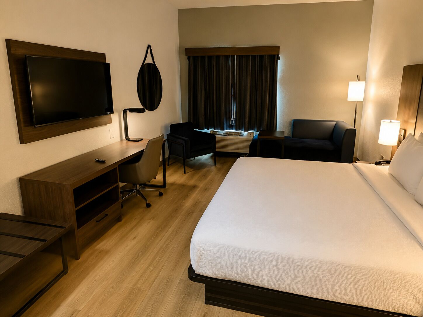 Une chambre d’hôtel avec un grand lit, un bureau avec une chaise, un téléviseur à écran plat fixé au mur, une lampe de bureau, une fenêtre avec des rideaux et une table de chevet avec une lampe.