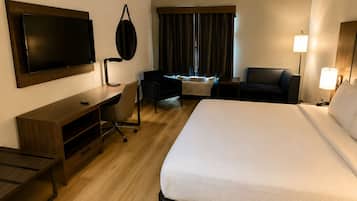 Ein Hotelzimmer mit einem großen Bett, einem Schreibtisch mit Stuhl, einem an der Wand befestigten Flachbildfernseher, einer Schreibtischlampe, einem Fenster mit Vorhängen und einem Nachttisch mit Lampe.