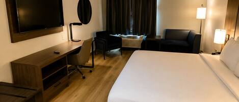 Habitación de hotel con una cama grande, un escritorio con una silla, un televisor de pantalla plana montado en la pared, una lámpara de escritorio, una ventana con cortinas y una mesita de noche con lámpara.