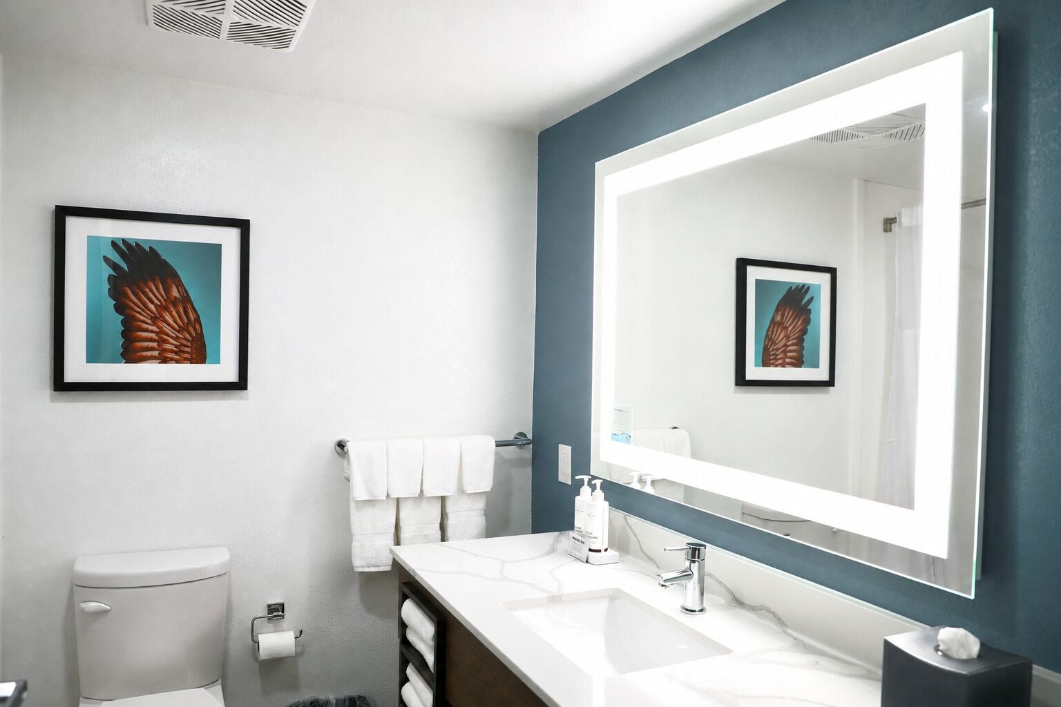 A modern bathroom with a large mirror, white sink, and a framed artwork on the wall.