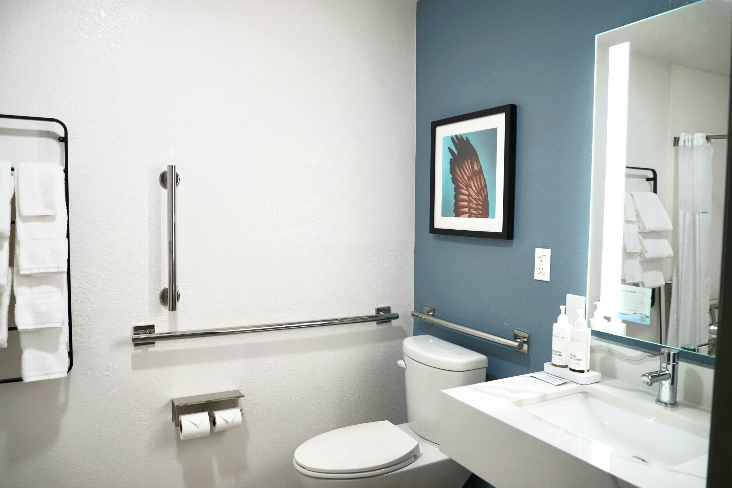 A bathroom with a white toilet, a sink, a mirror, a towel rack, and a framed picture on the wall.