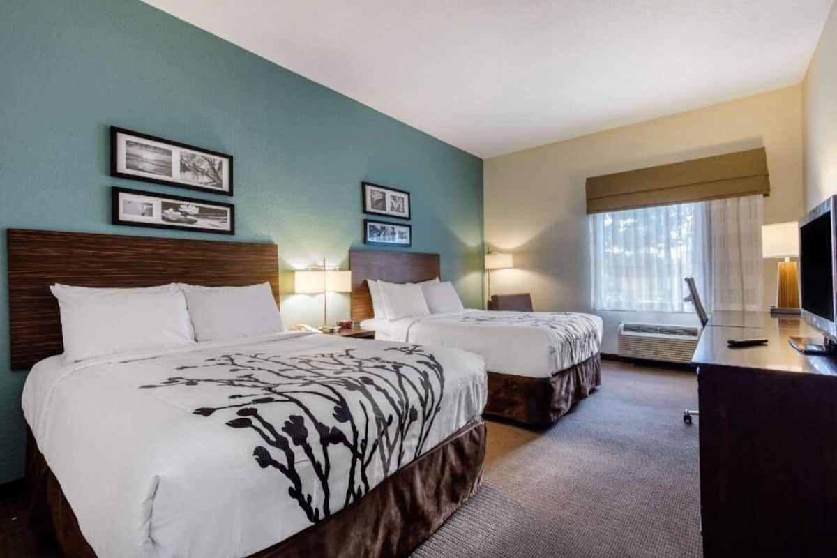 A hotel room with two beds, a desk, a TV, and framed pictures on the wall.