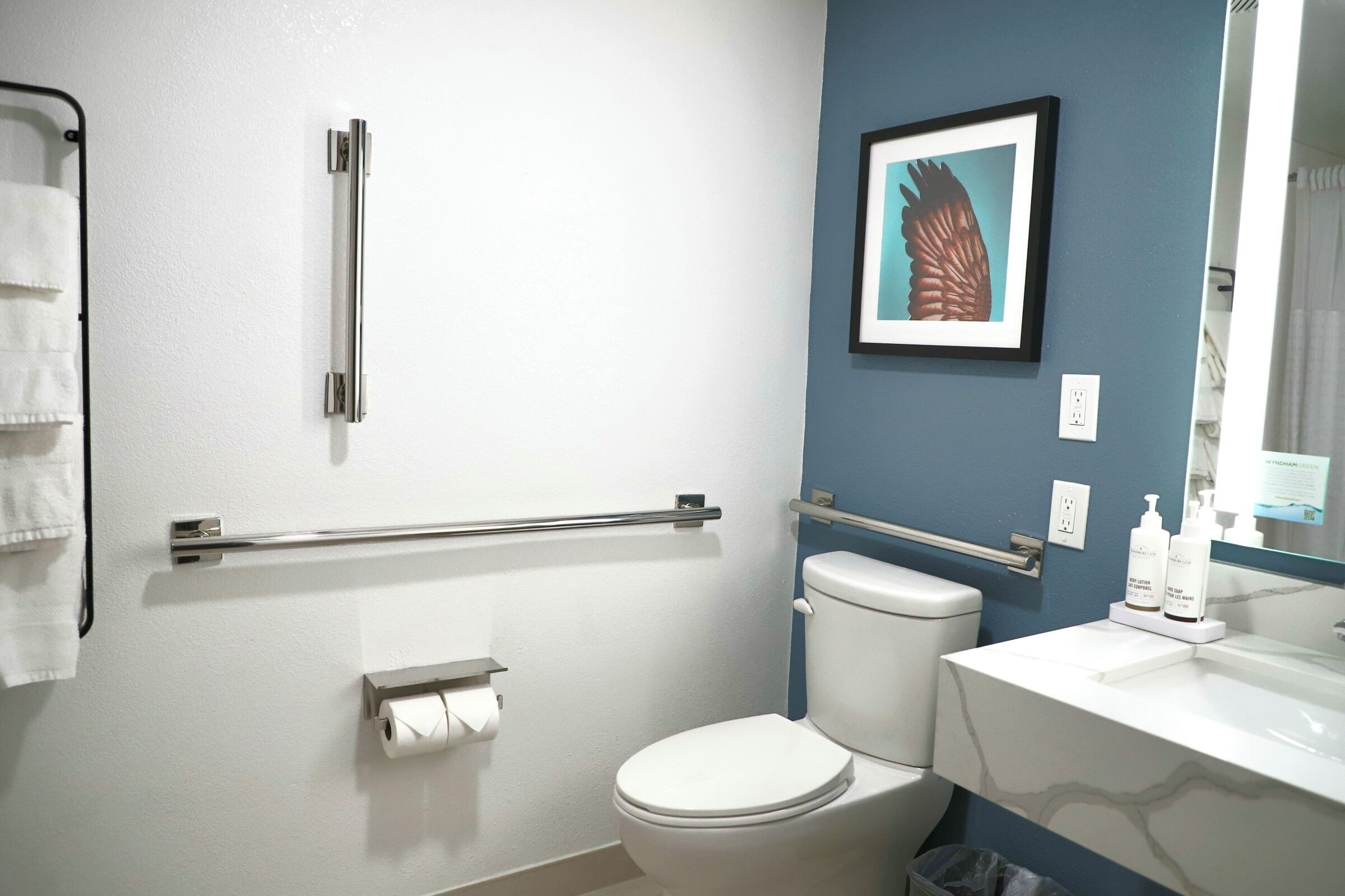 A bathroom with a white toilet, a towel rack, a handrail, a framed picture on the wall, and a sink with toiletries.