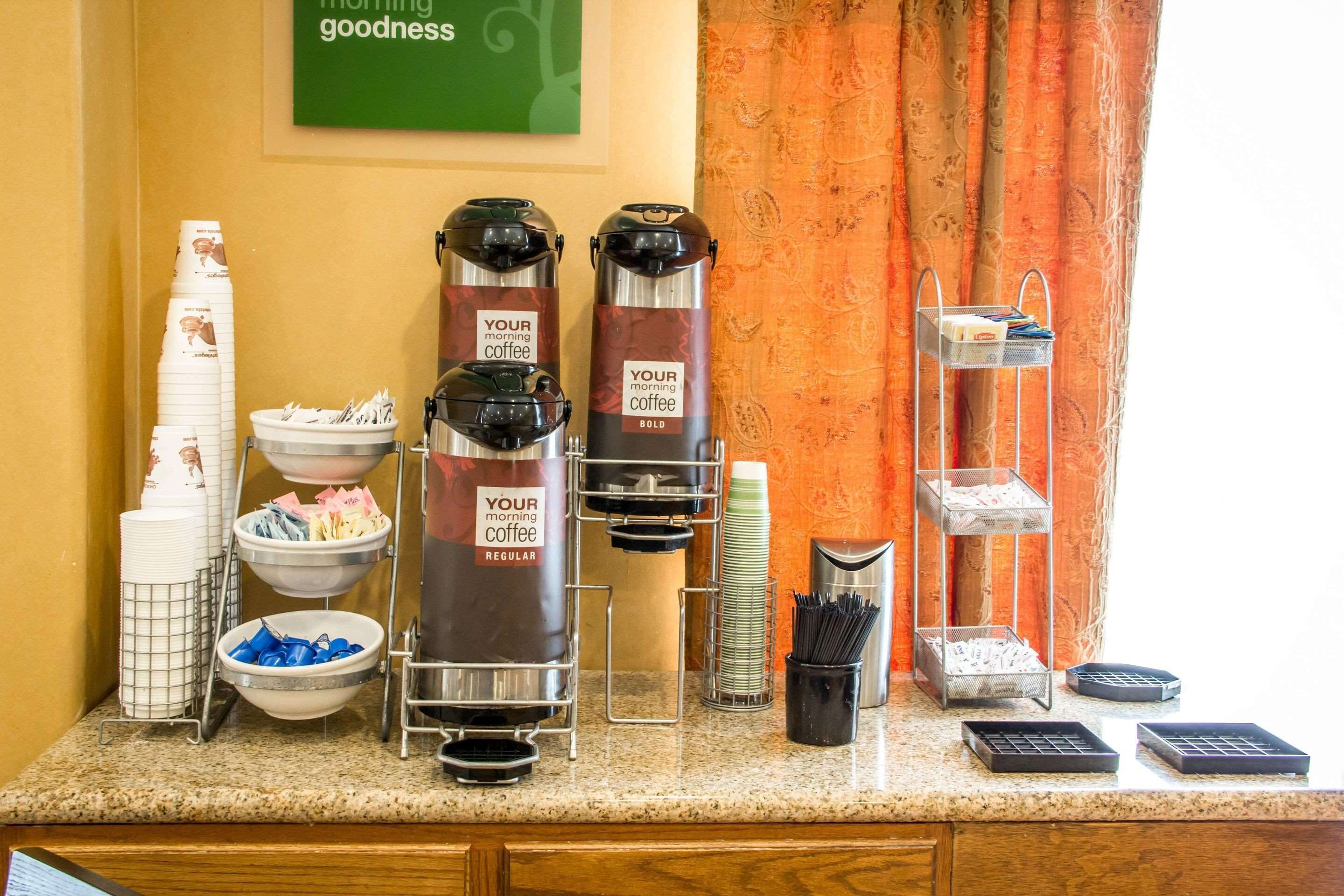 A coffee station with two coffee dispensers labeled "Your morning coffee" and "Your morning coffee bold," along with various coffee cups, a bowl of utensils, and a tray of newspapers.