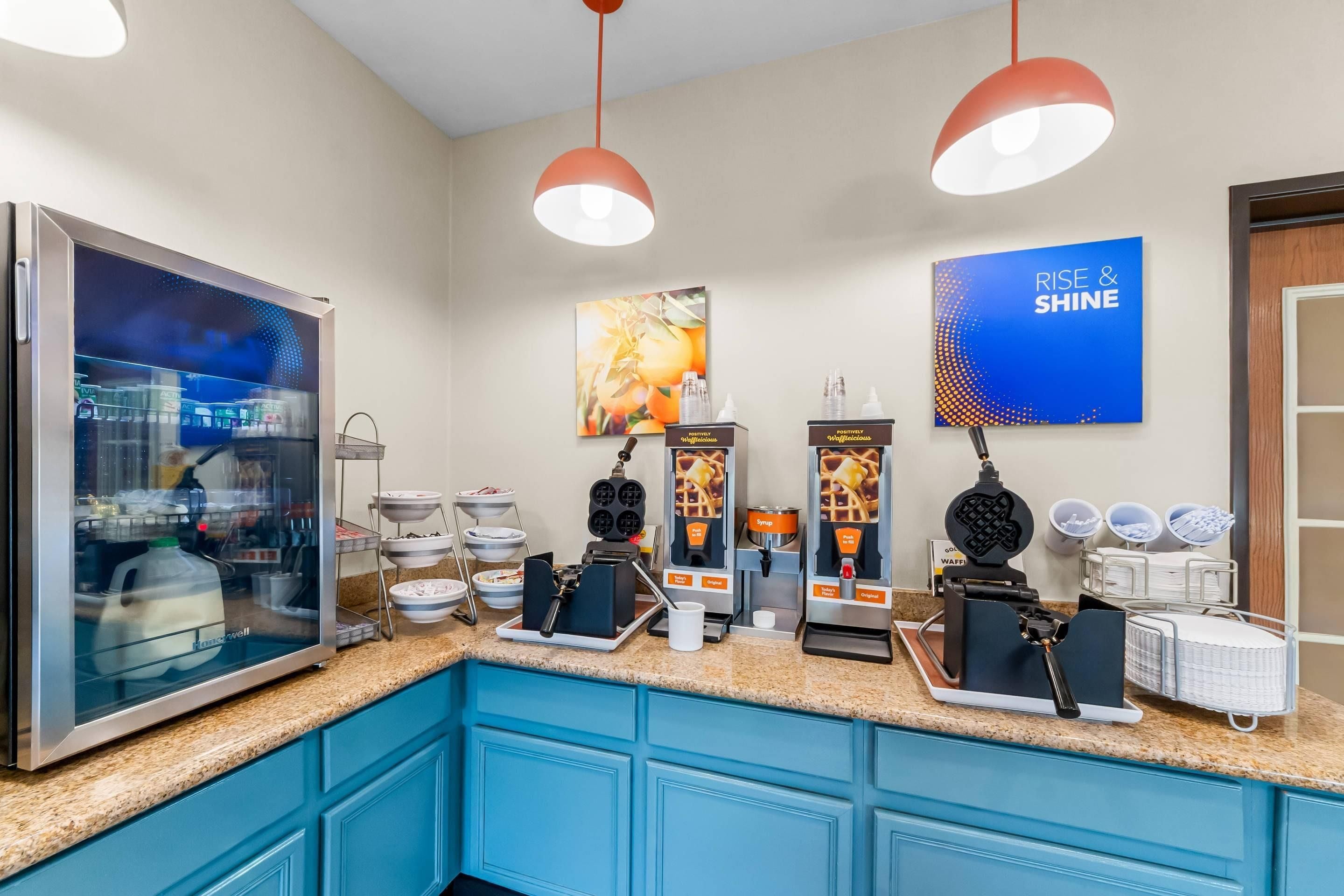 A self-service breakfast area with waffle makers, syrup dispensers, and a refrigerator.