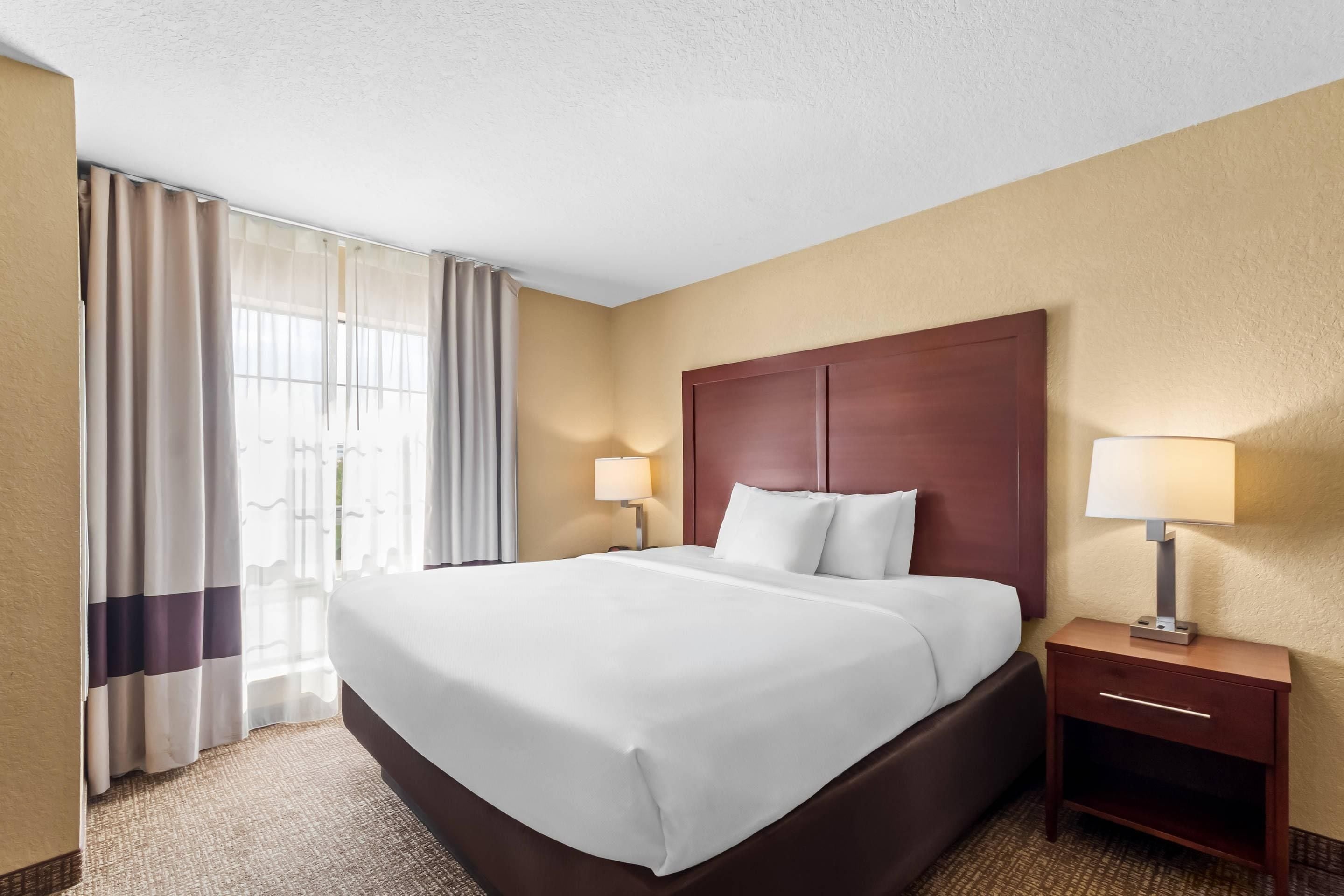 A hotel room with a large bed, two bedside tables with lamps, a window with curtains, and a carpeted floor.