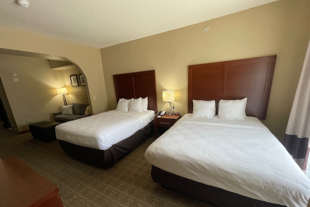 Habitación de hotel con dos camas individuales y una cama doble, un escritorio con lámpara y una alfombra estampada.