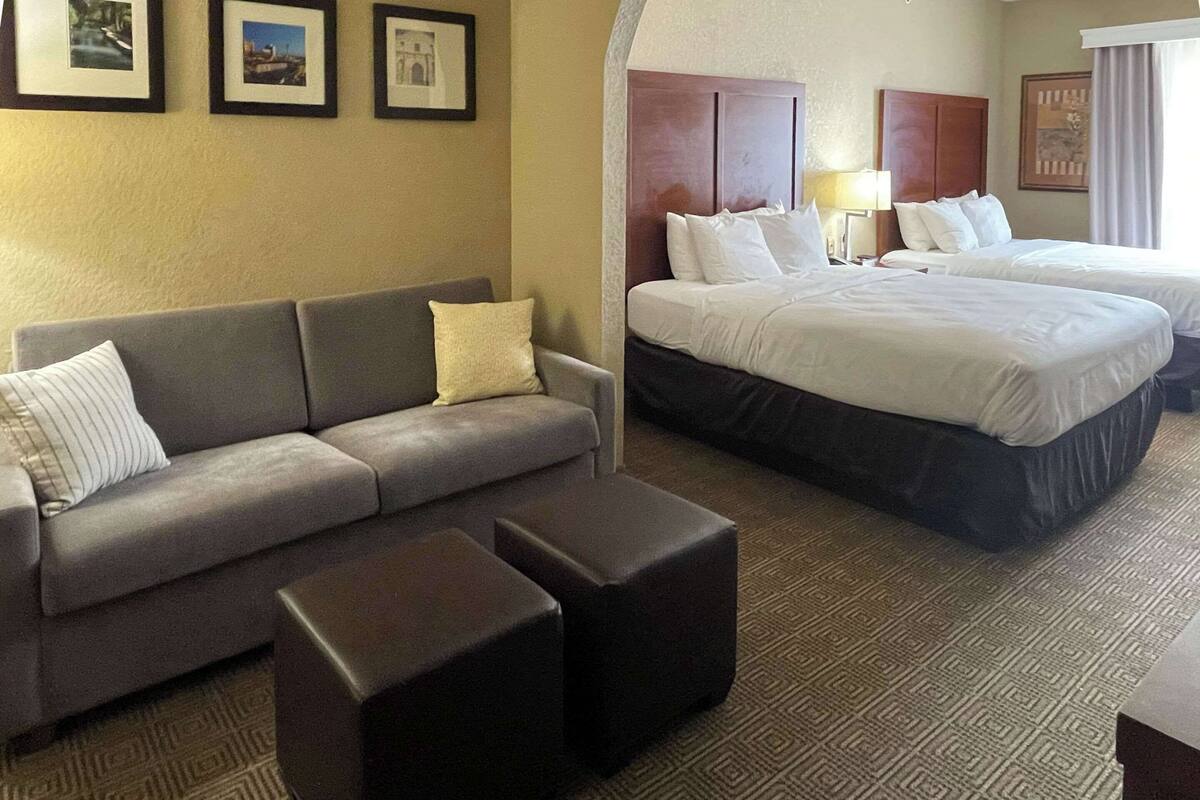 Una camera d'albergo con un divano grigio, due letti e un tappeto decorato.