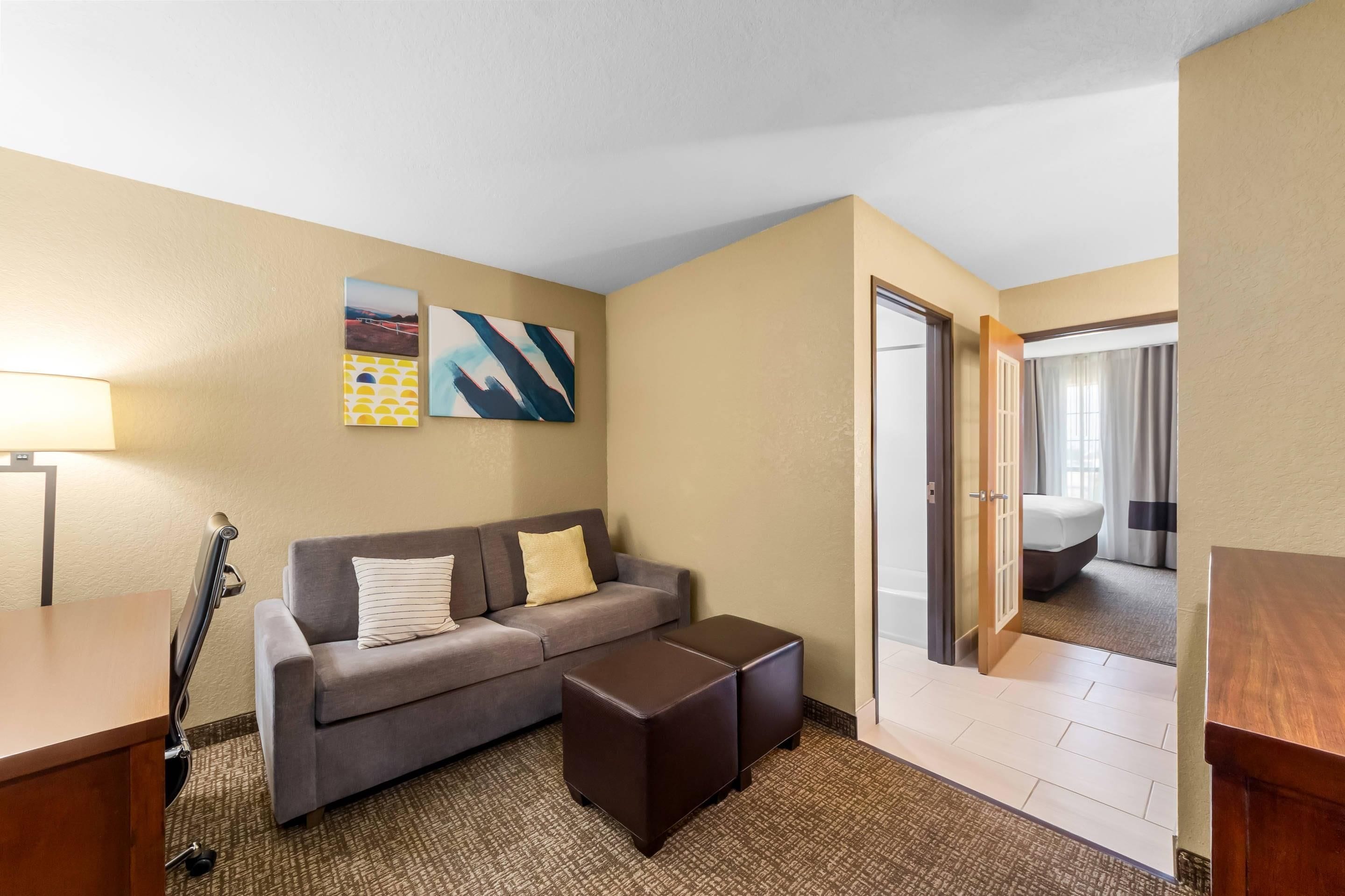 Una habitación de hotel con sofá, reposapiés, lámpara y una obra de arte en la pared.
