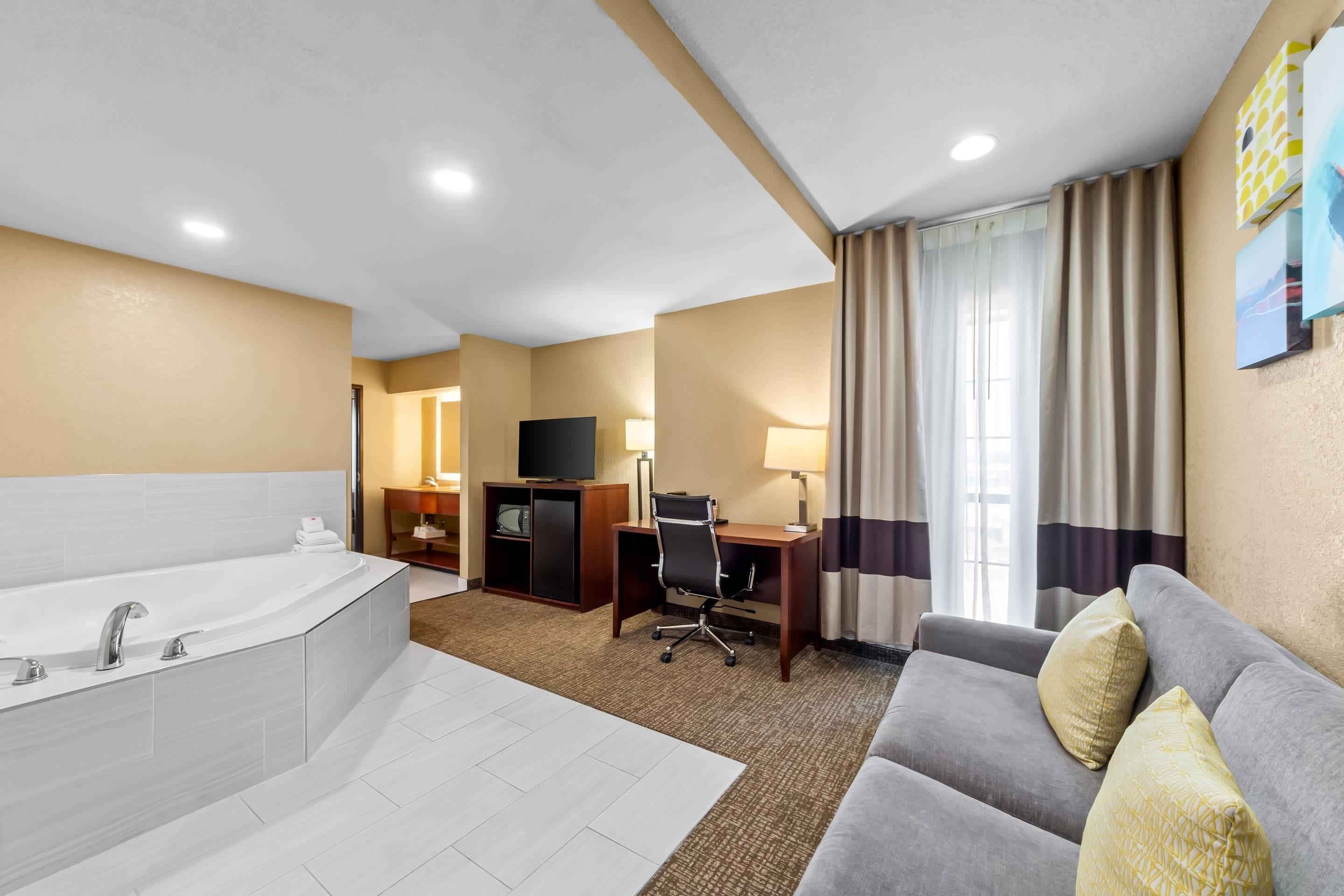 A hotel room with a large bathtub, a desk with a chair, a television, and a sofa with pillows.