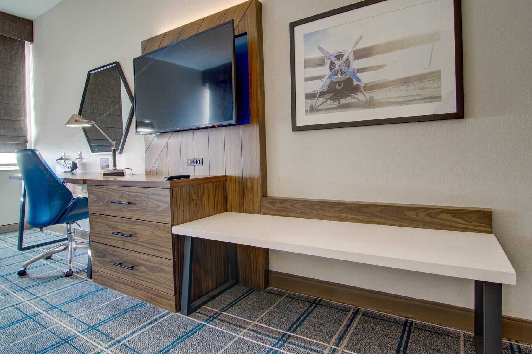 A hotel room with a desk, a chair, a flat-screen TV, a framed picture, and a patterned carpet.