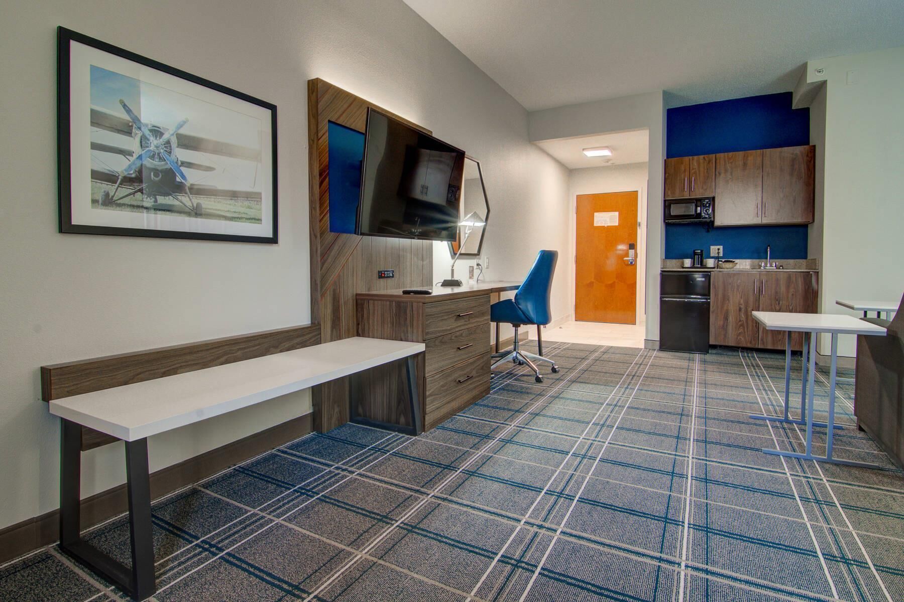 Habitación de hotel con una banca, un escritorio, una silla, un televisor, una kitchenette y una alfombra estampada.