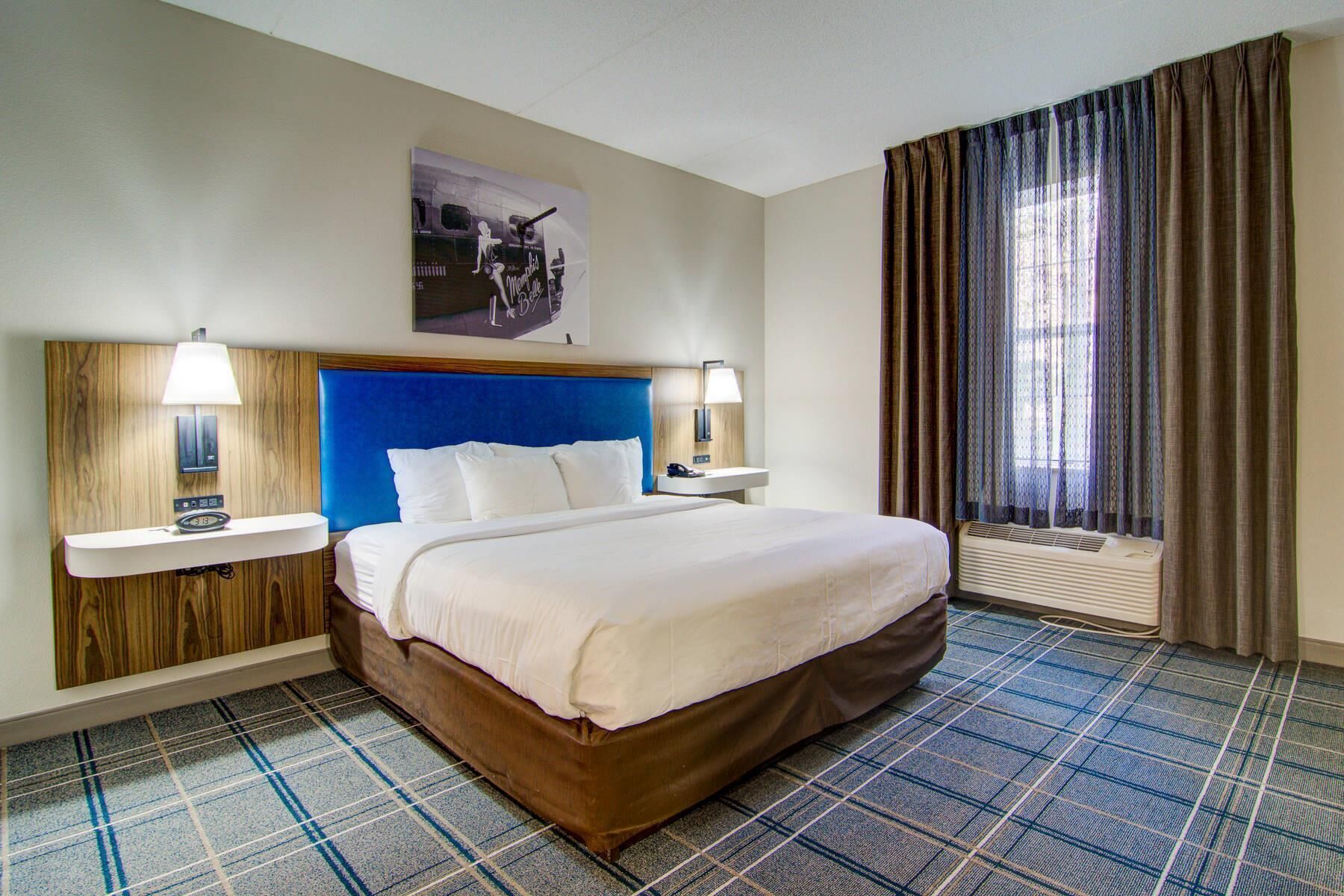 Une chambre d’hôtel comprenant un lit, deux tables de chevet, une petite table, une horloge murale, une fenêtre avec des rideaux et un climatiseur fixé au mur.