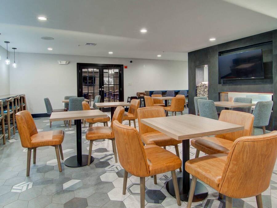 Een modern restaurant met een vloer van hexagonaal patroon, houten tafels en stoelen, inbouwspots en een groot flatscreen televisietoestel aan de muur.